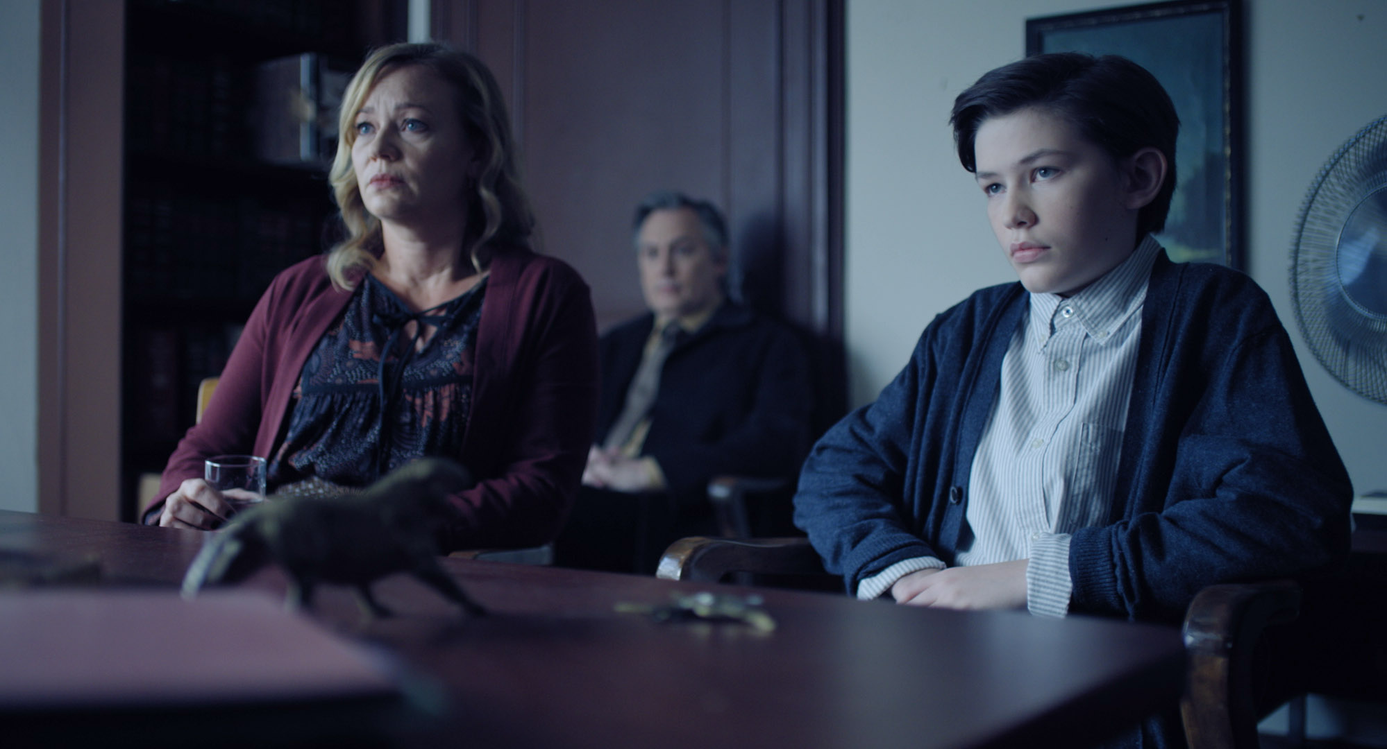 Jacob and his mother sit at a desk looking concerned. There is a figurine of an animal on the desk in the foreground and a man sitting in the background.