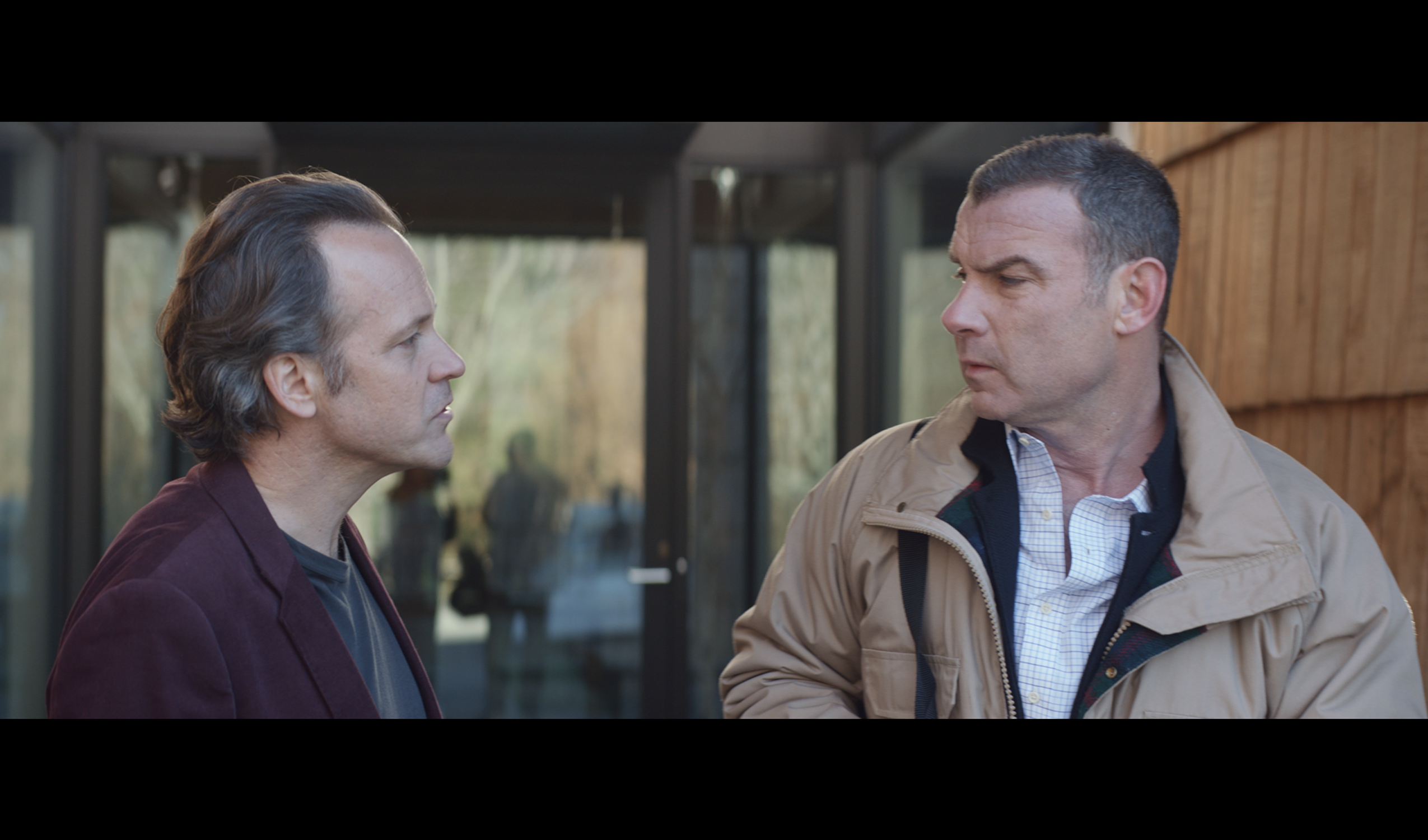 A still from the film, where two middle-aged white men face each other in front of a glass wall. One is frowning slightly and they look as if they are in the middle of a conversation.