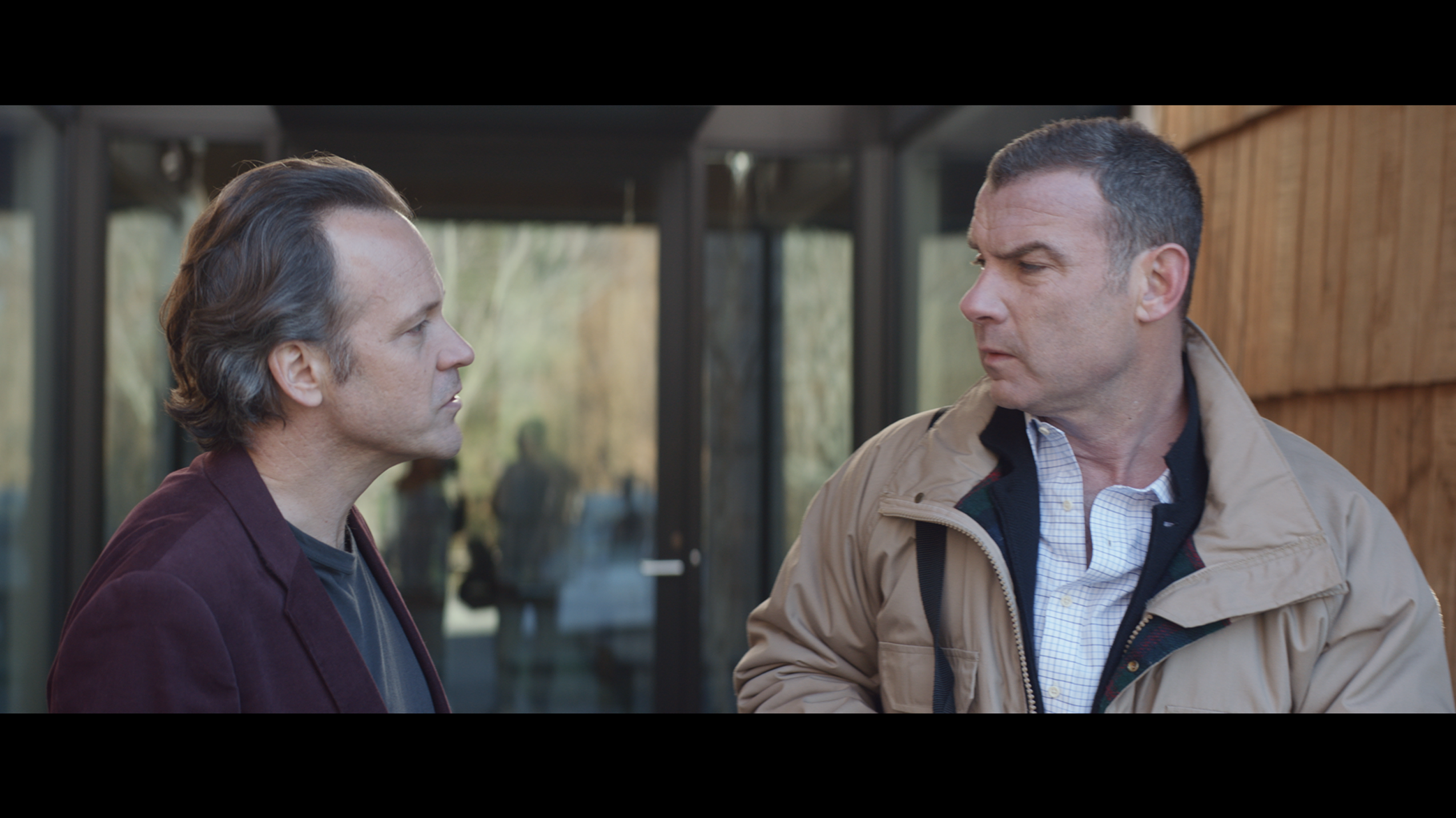 A still from the film, where two middle-aged white men face each other in front of a glass wall. One is frowning slightly and they look as if they are in the middle of a conversation.