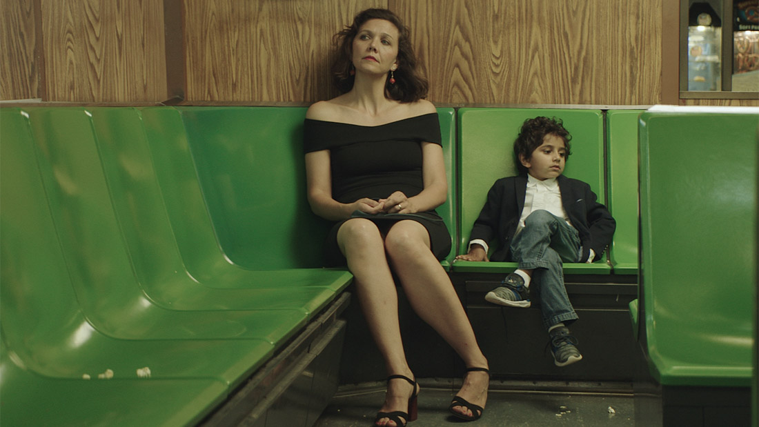 Maggie Gyllenhaal wears a black dress and is looking into the distance to the left. A small child is sitting next to her with his legs crossed, looking in the opposite direction. The chairs are green plastic and the walls are a medium-tone wood with prominent grain.