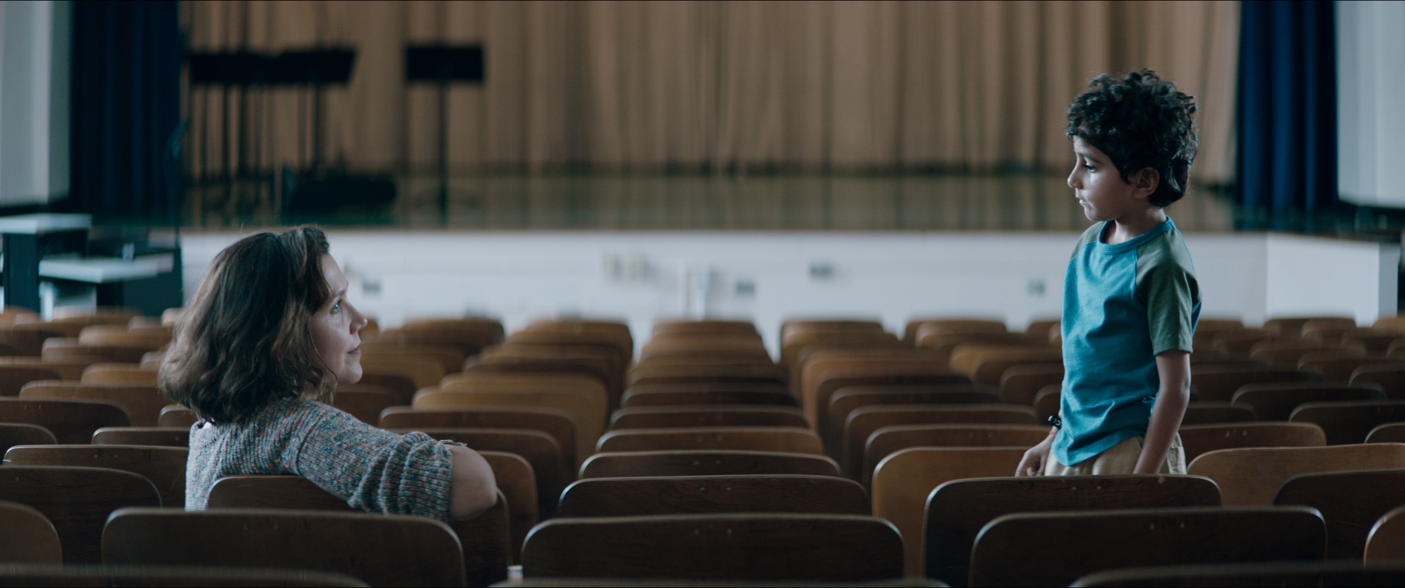 Maggie Gyllenhaal is seated in an auditorium, facing Parker Sevak, who is standing wearing a blue shirt and looking back at Maggie. The seats are brown and in the background is a stage with a closed brown curtain and some music stands.