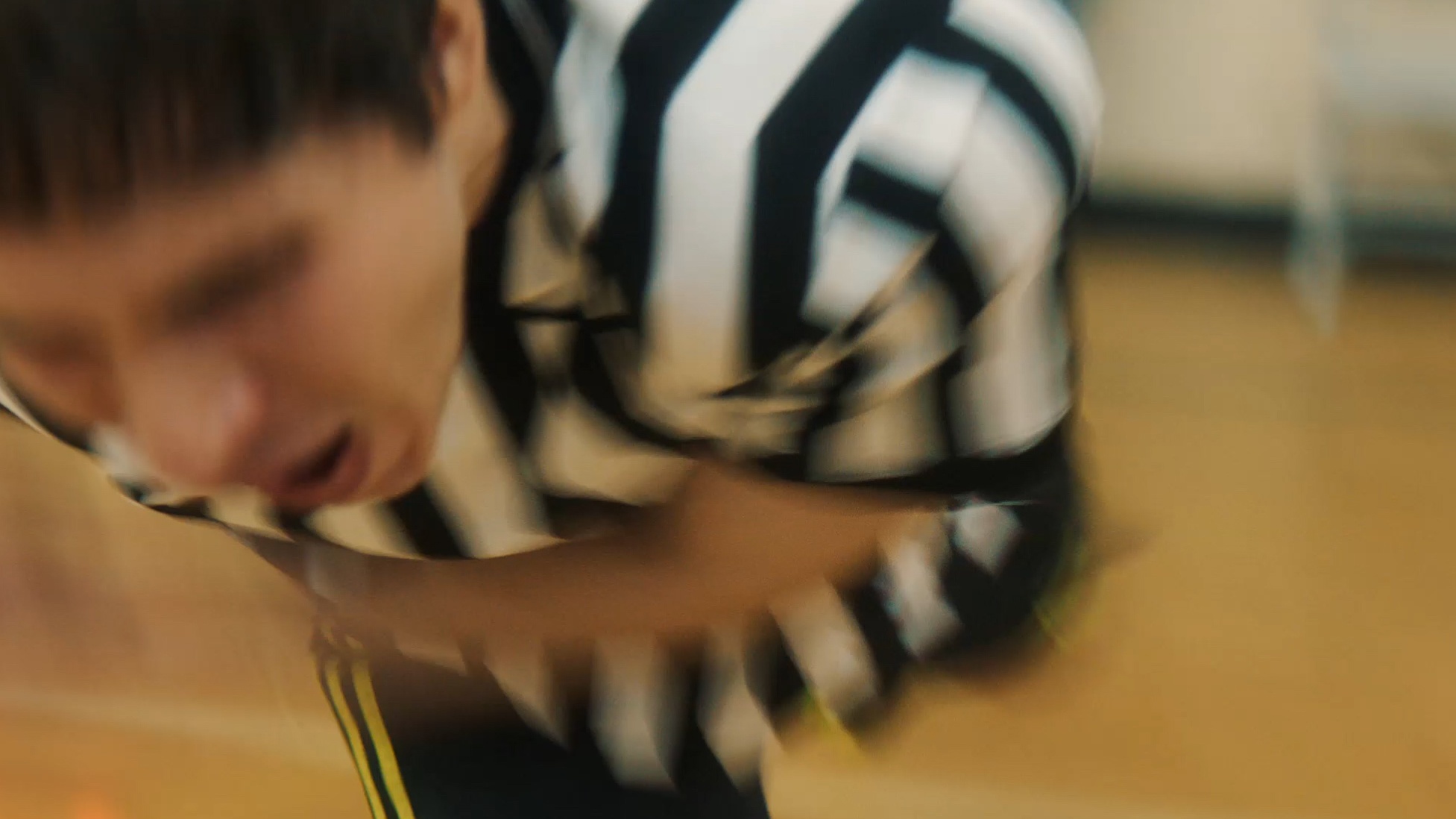 A blurry capture of a person in a referee's black-and-white striped jersey stumbling forward, mouth open, with a wooden gym floor in the background.
